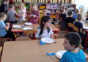 Dzieci siedzące przy stolikach i krzątające się między stolikami podczas nakrywania do stołu.